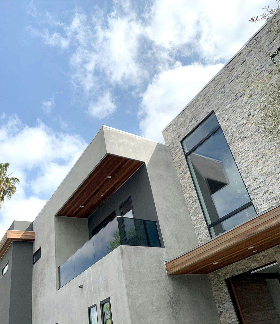 A photograph of the exterior facade of the home chosen for the event venue. On one side the facade is cement and has a balcony. The other side is a stacked slate. Both facades are square and have wooden and steel accents and large windows.