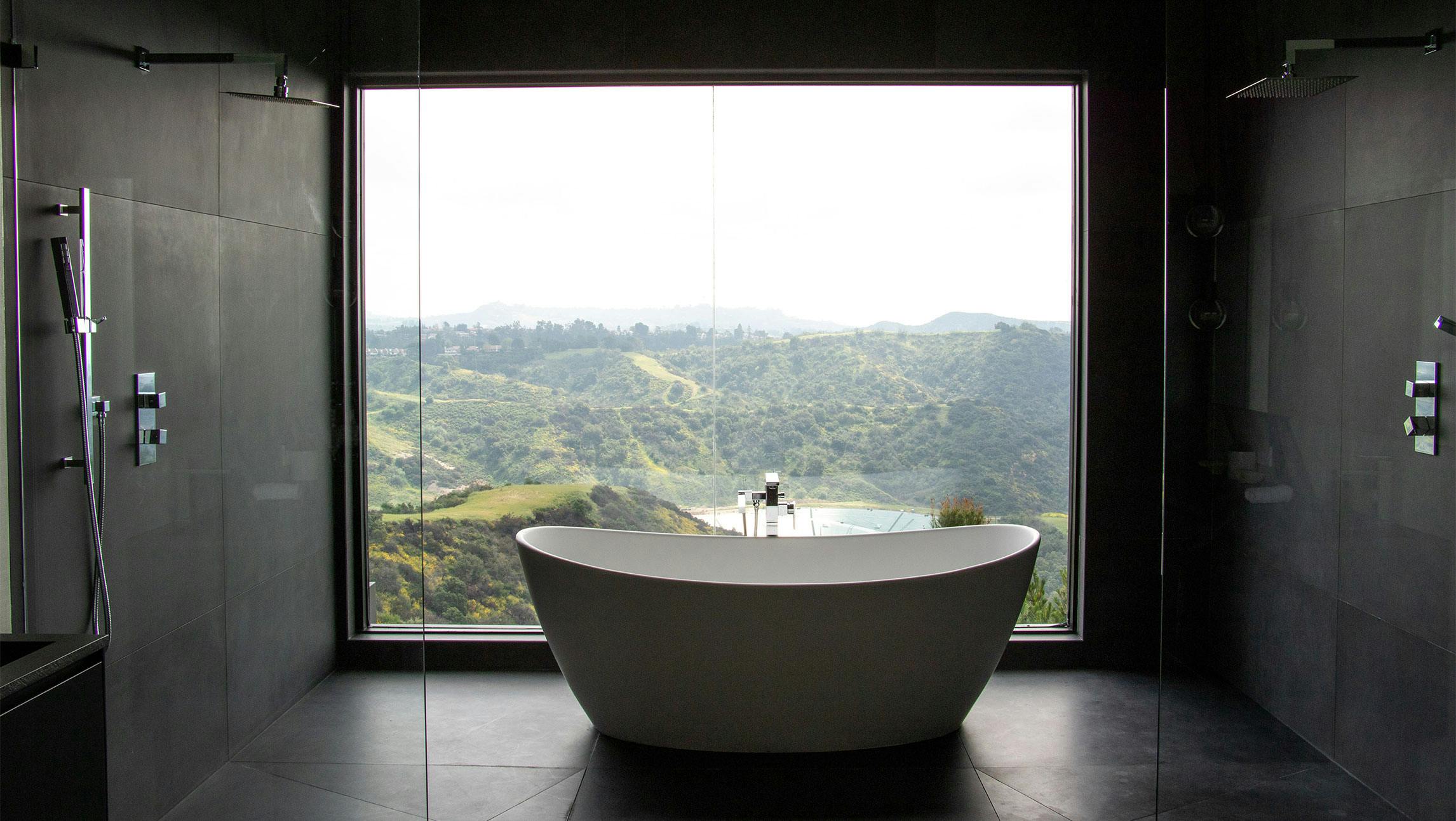 A photograph of a bathtub with a view. The white tub is centered in a dark slate room with chrome faucets and one wall that is all glass and overlook the green hills outside.