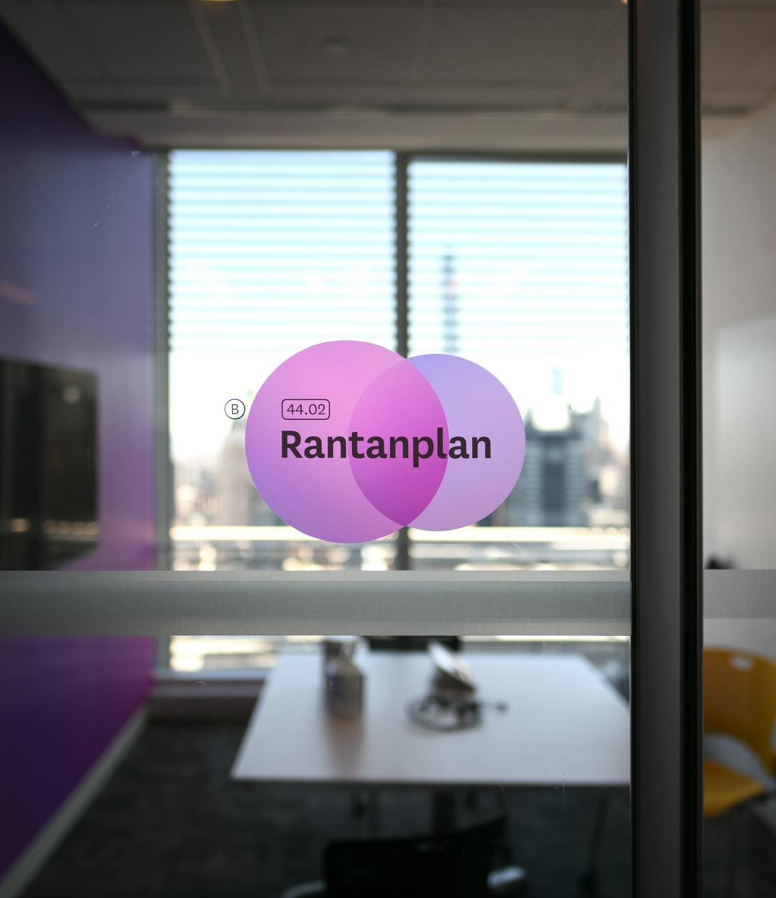 A close up photograph of a conference room sign adhered to the glass wall of a Datadog conference room. The sign is comprised of two purple circles, one overlapping the other, with the conference room name, 'Rantanplan' in bold lettering on top of the layered shape.