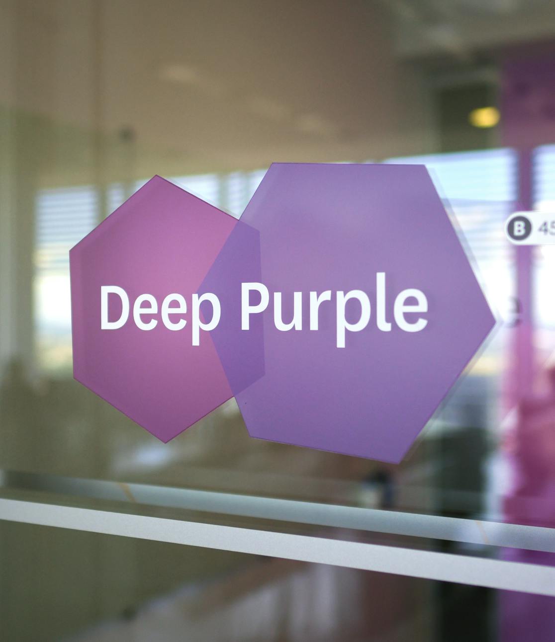 A close up photograph of a conference room sign adhered to the glass wall of a Datadog conference room. The sign is comprised of two purple hexagons, one overlapping the other, with the conference room name, 'Deep Purple' in bold lettering on top of the layered shape.