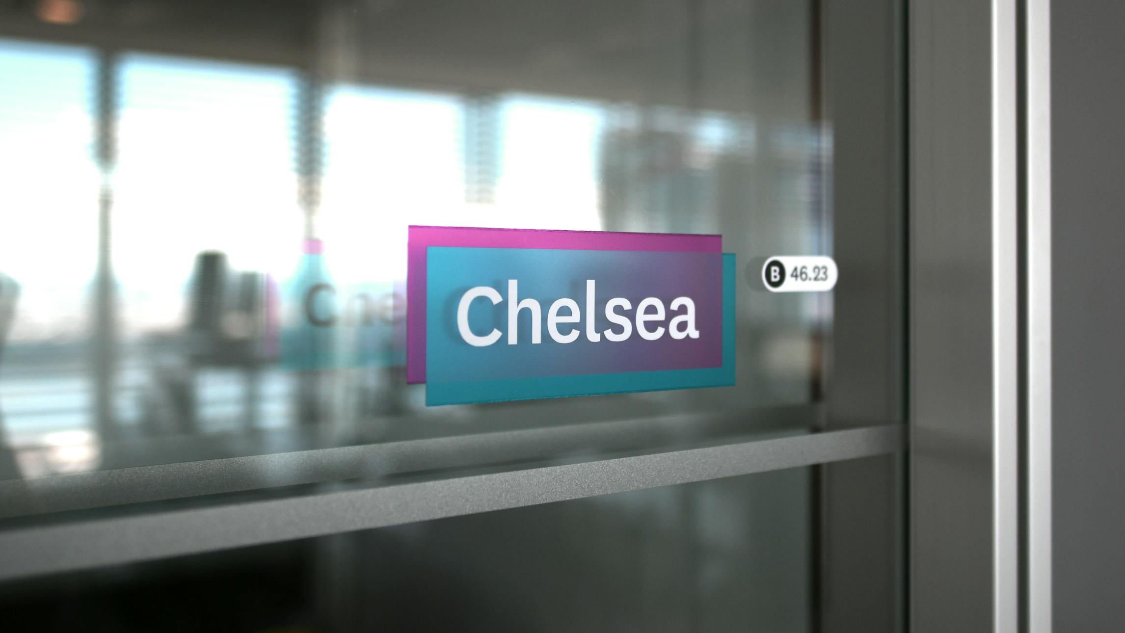 A close up photograph of a conference room sign adhered to the glass wall of a Datadog conference room. The sign is comprised of a blue rectangle overlapping a purple rectangle with the conference room name, 'Chelsea' in bold lettering on top of the layered shape.