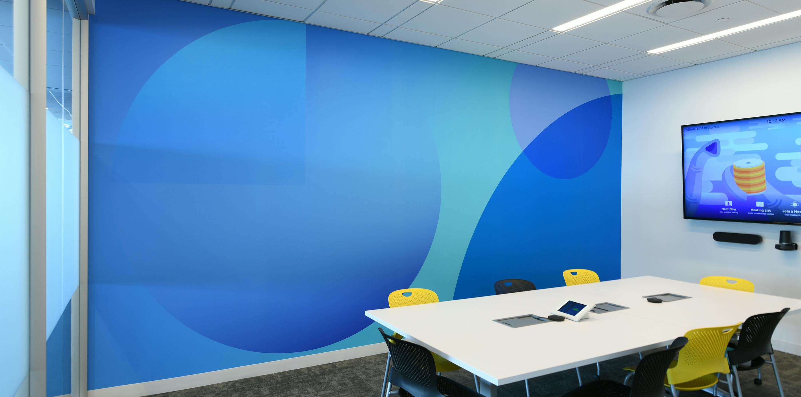 A photograph of a Datadog conference room contaning a white table with eight chairs(four yellow and four black), a television and video camera mounted on one wall and an abstract monochromatic blue wallpaper on another wall.