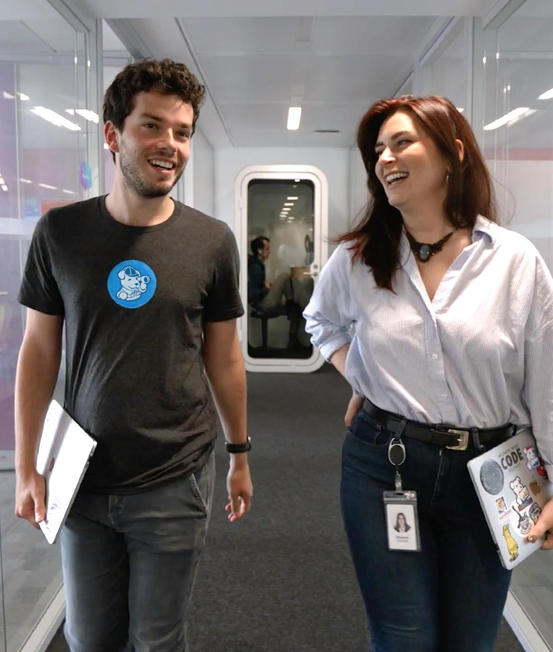 A thumbnail image taken from a Datadog recruiting video; Two colleagues dressed in casual attire and walking by conference rooms in a Datadog office.